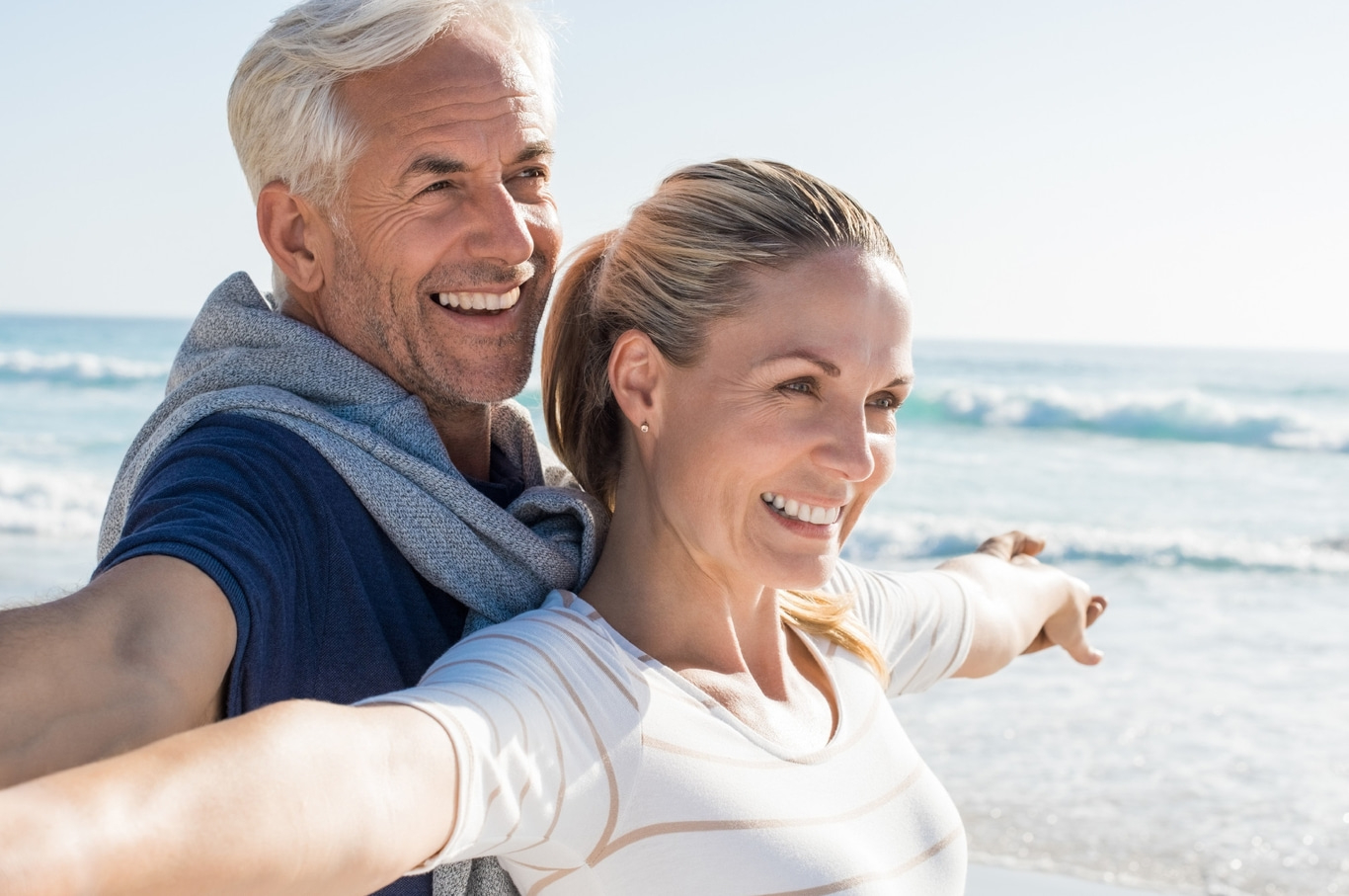 Pareja feliz en la playa, brazos extendidos, sonriendo, día soleado, mar de fondo.